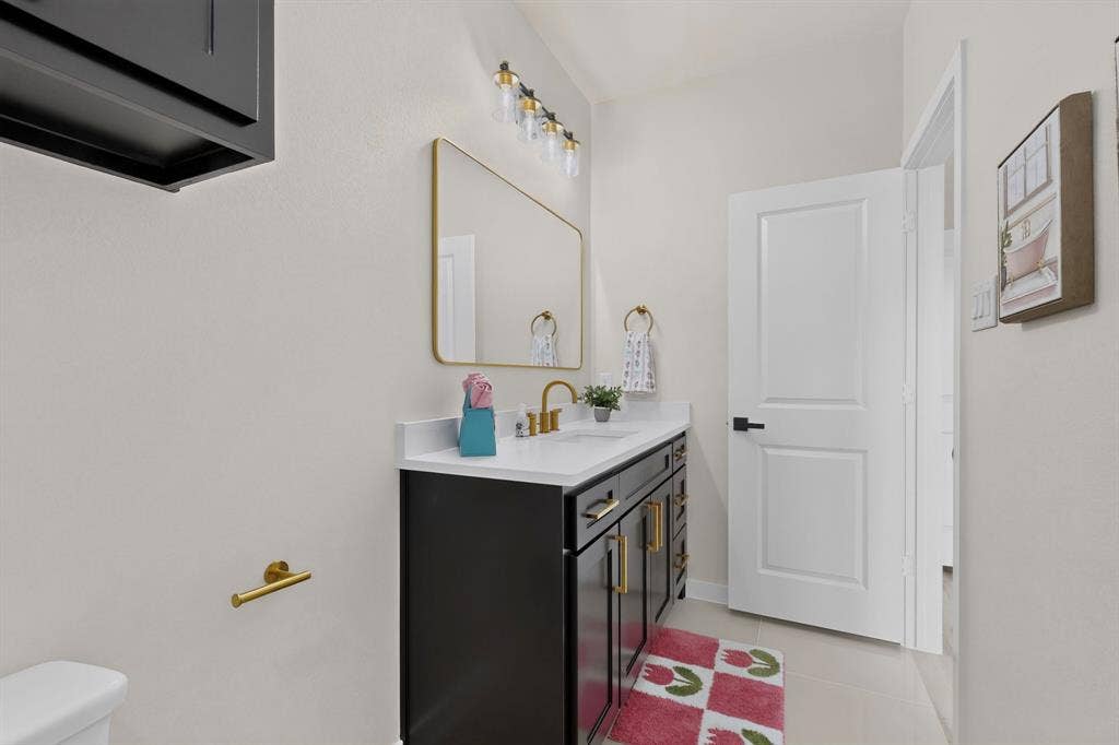 102 Nicklaus Drive, Sulphur Springs, TX 75482 Vanity featuring dark cabinetry, white countertop, gold-toned hardware, rectangular mirror, and multi-bulb wall-mounted lighting