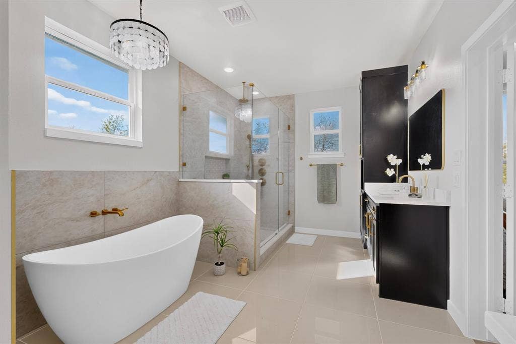 102 Nicklaus Drive, Sulphur Springs, TX 75482 Freestanding soaking tub with wall-mounted gold-toned faucet, glass-enclosed shower with tiled surround, dual-toned cabinetry, light-toned tile flooring, and decorative crystal chandelier