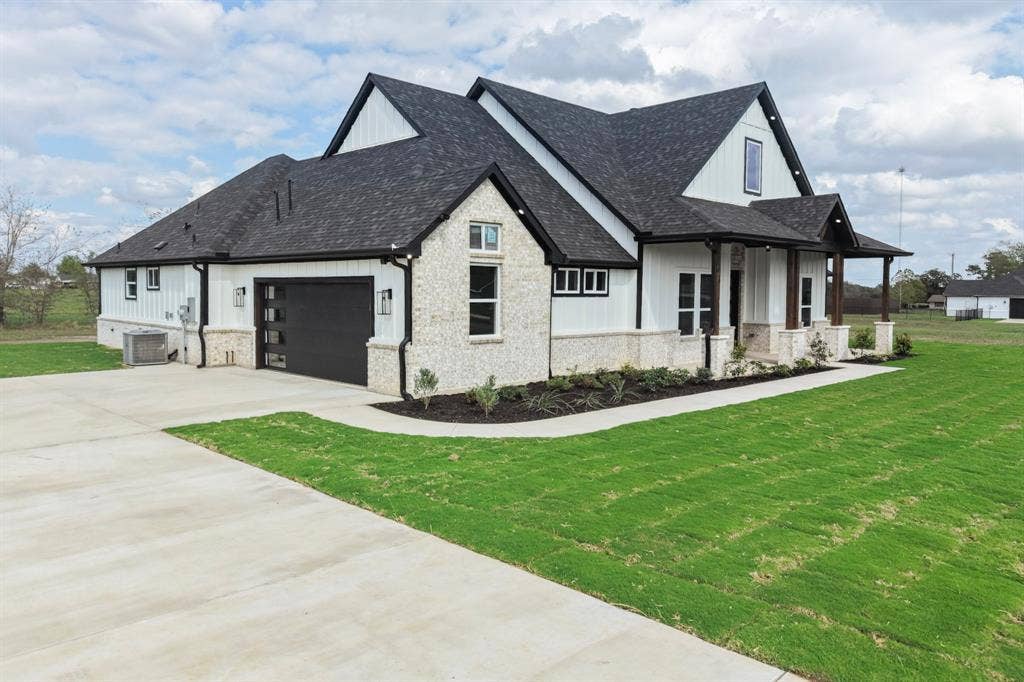 102 Nicklaus Drive, Sulphur Springs, TX 75482 Modern farmhouse exterior featuring vertical board-and-batten siding, stone masonry accents, a steep gabled roofline, and a multi-panel garage door