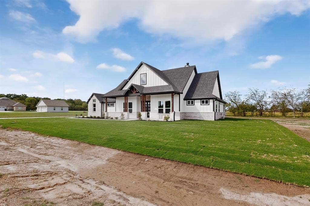 102 Nicklaus Drive, Sulphur Springs, TX 75482 Modern farmhouse exterior featuring vertical white siding, dark shingled roof, stone masonry accents, wood-beam porch supports, and a dedicated detached garage