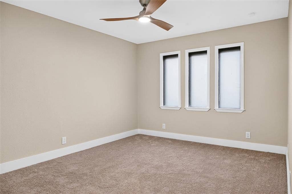 Neutral-toned interior room featuring wall-to-wall carpeting, three vertical windows, and a ceiling-mounted light fixture with wood-finish blades