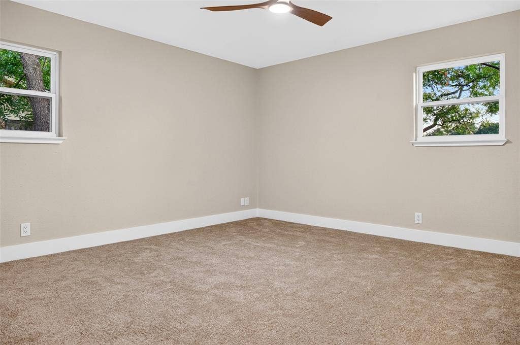 Neutral-toned interior featuring wall-to-wall carpeting, dual operable windows with exterior foliage, and a ceiling-mounted fan with integrated lighting