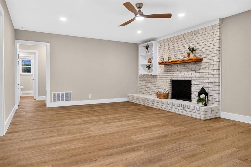 Painted brick fireplace with a wood mantel, wood-finish flooring, built-in shelving, and a modern ceiling fan