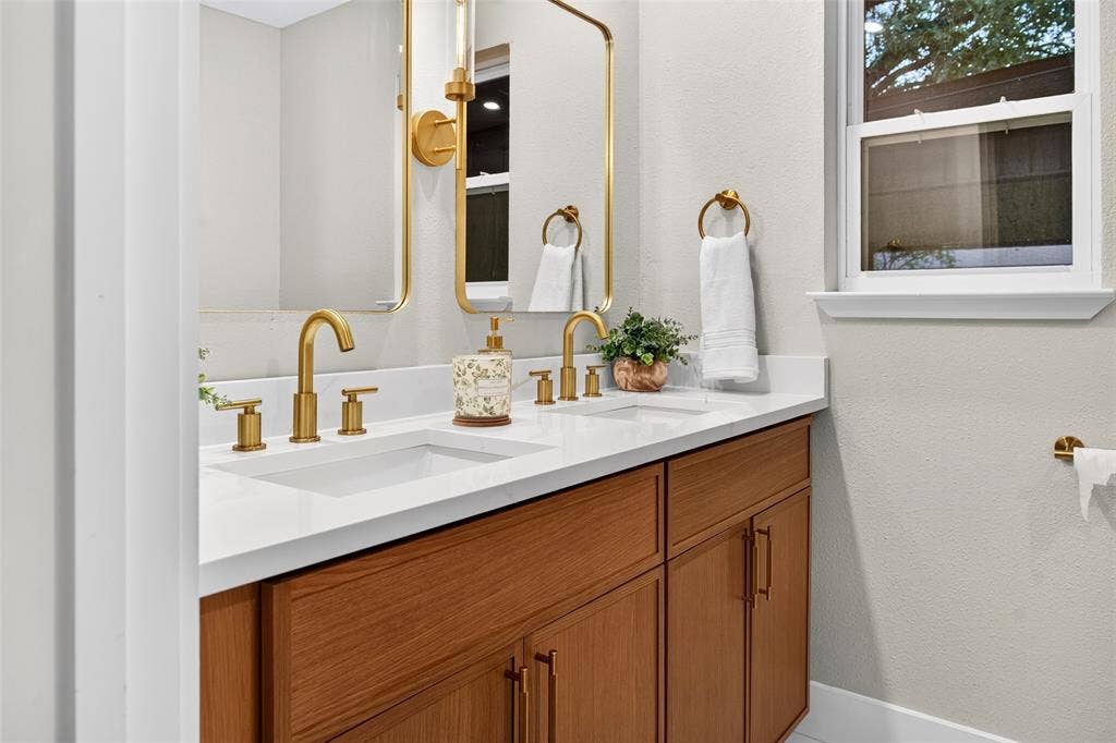 Dual-sink vanity featuring wood-finish cabinetry, quartz countertop, polished brass hardware, and rectangular mirrors
