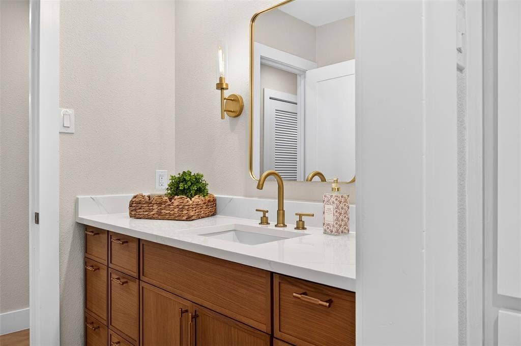 Wood-finish cabinetry, quartz countertop, rectangular undermount sink, brass-finish widespread faucet, and matching brass-rimmed mirror