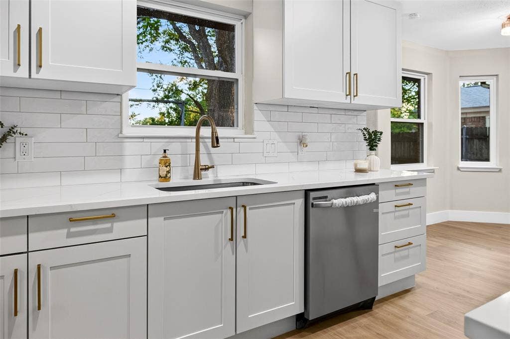Kitchen featuring white Shaker-style cabinetry, quartz countertops, subway tile backsplash, stainless steel dishwasher, and gold-finish hardware