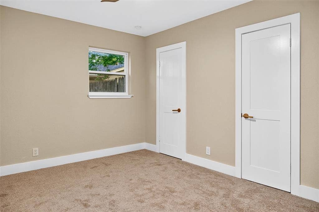 Neutral-toned interior room featuring wall-to-wall carpeting, two white shaker-style doors with metallic hardware, and a single-pane window
