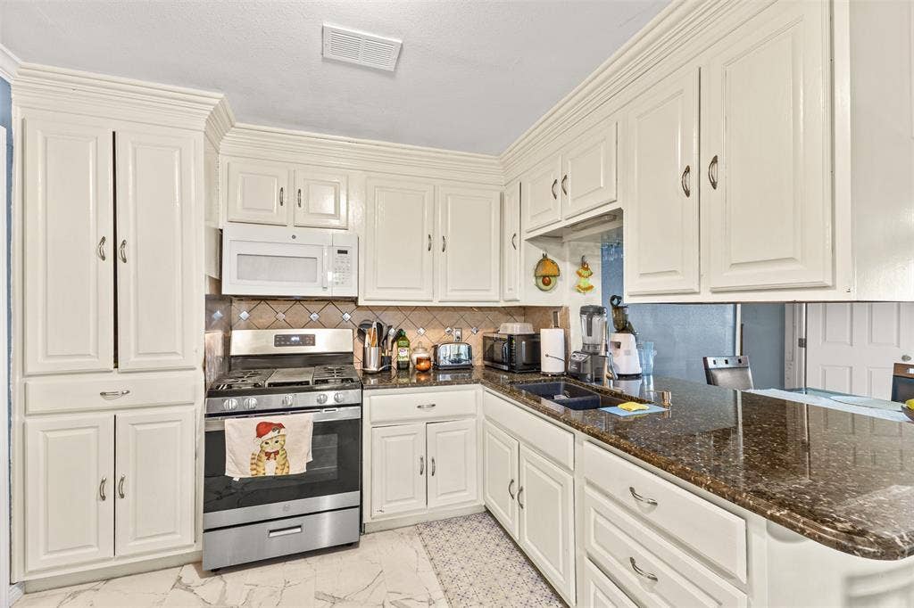 1801 Pleasant Run Road, Carrollton, TX 75006 Kitchen featuring off-white cabinetry with crown molding, dark stone-finish countertops, stainless steel gas range, over-the-range microwave, and stone tile flooring