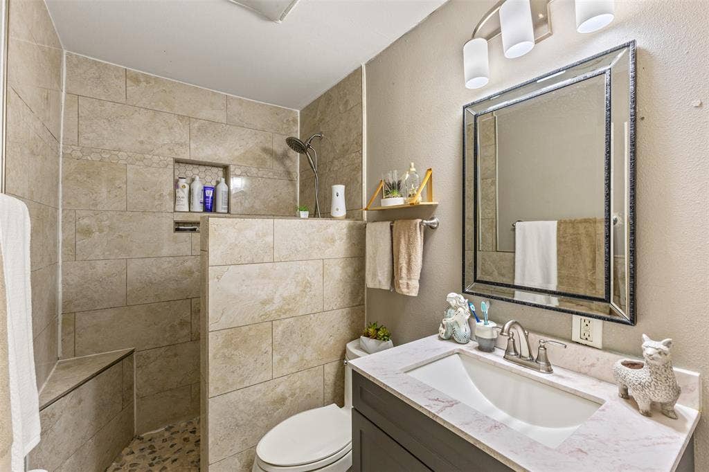 1801 Pleasant Run Road, Carrollton, TX 75006 Walk-in shower with stone tile surround, built-in bench, and pebble flooring, featuring a dark-finish vanity with a white countertop and rectangular undermount sink