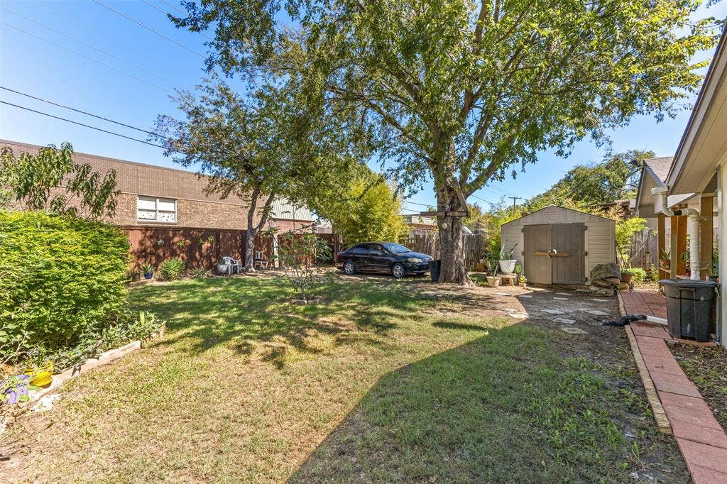 1801 Pleasant Run Road, Carrollton, TX 75006 Backyard area featuring a manicured lawn, mature trees, a detached storage shed, and a brick-paver pathway