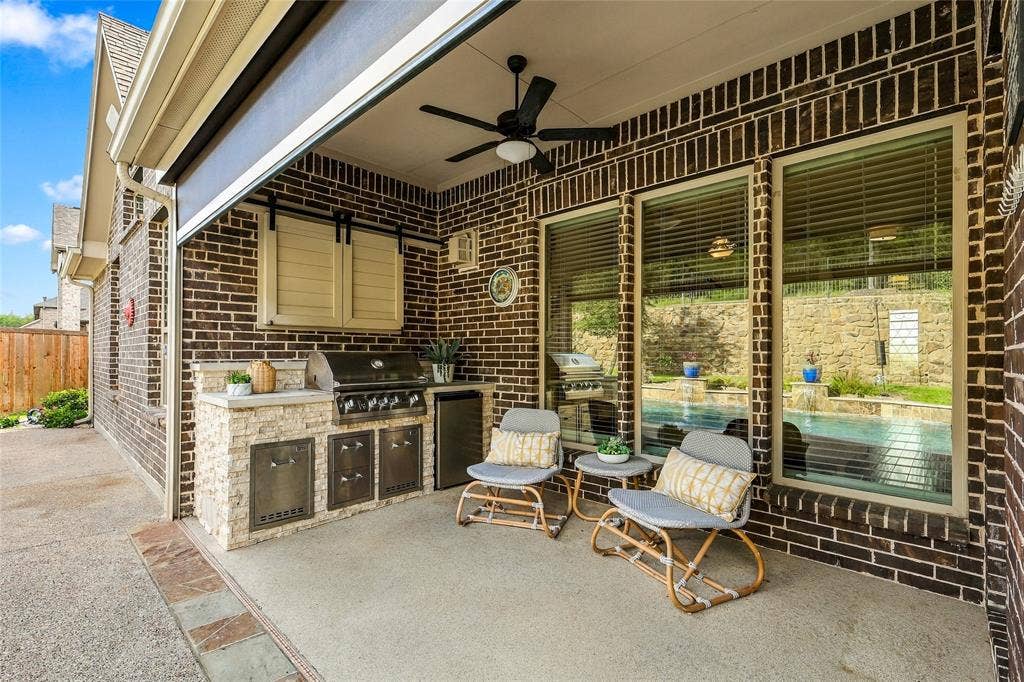 Covered patio featuring brick masonry, a built-in outdoor kitchen with stainless steel appliances, stone-veneer cabinetry, ceiling fan, and remote contolled patio screen