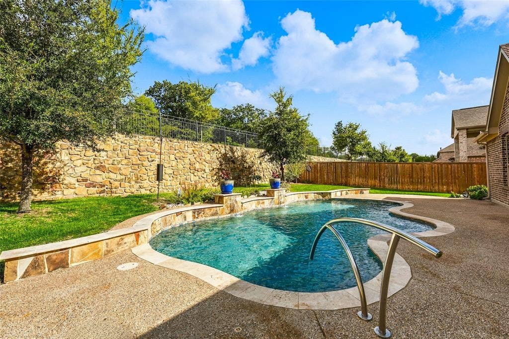 Free-form swimming pool with stone coping, decorative water feature, aggregate patio surface, and stone retaining wall
