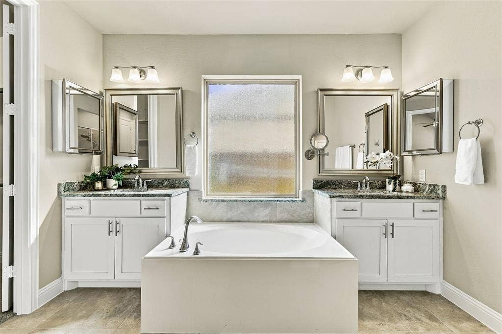 Dual white vanities featuring stone-finish countertops, undermount sinks, framed mirrors, and central soaking tub with an arched faucet and frosted glass window