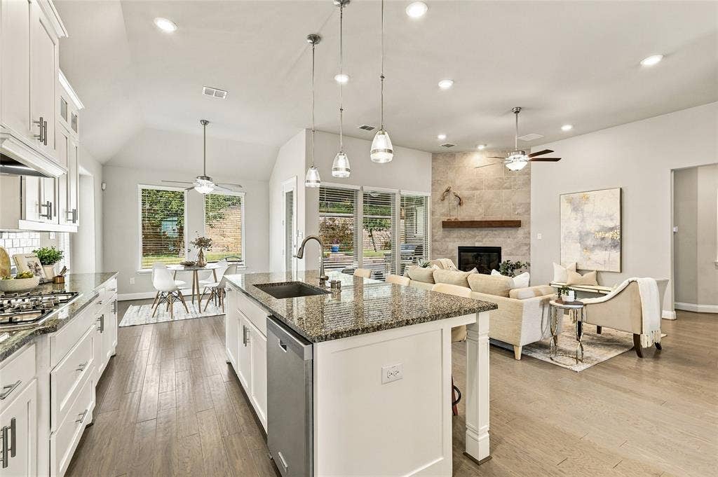 Open-concept kitchen and living area featuring wood-finish flooring, a central island with stone countertops, vaulted ceilings, pendant lighting, and a floor-to-ceiling stone fireplace surround with a wood mantel