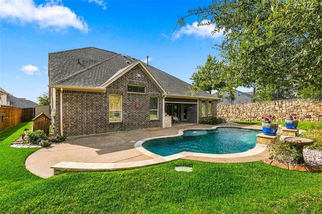 Brick exterior home featuring a rear swimming pool, concrete patio, stone retaining wall, and manicured lawn