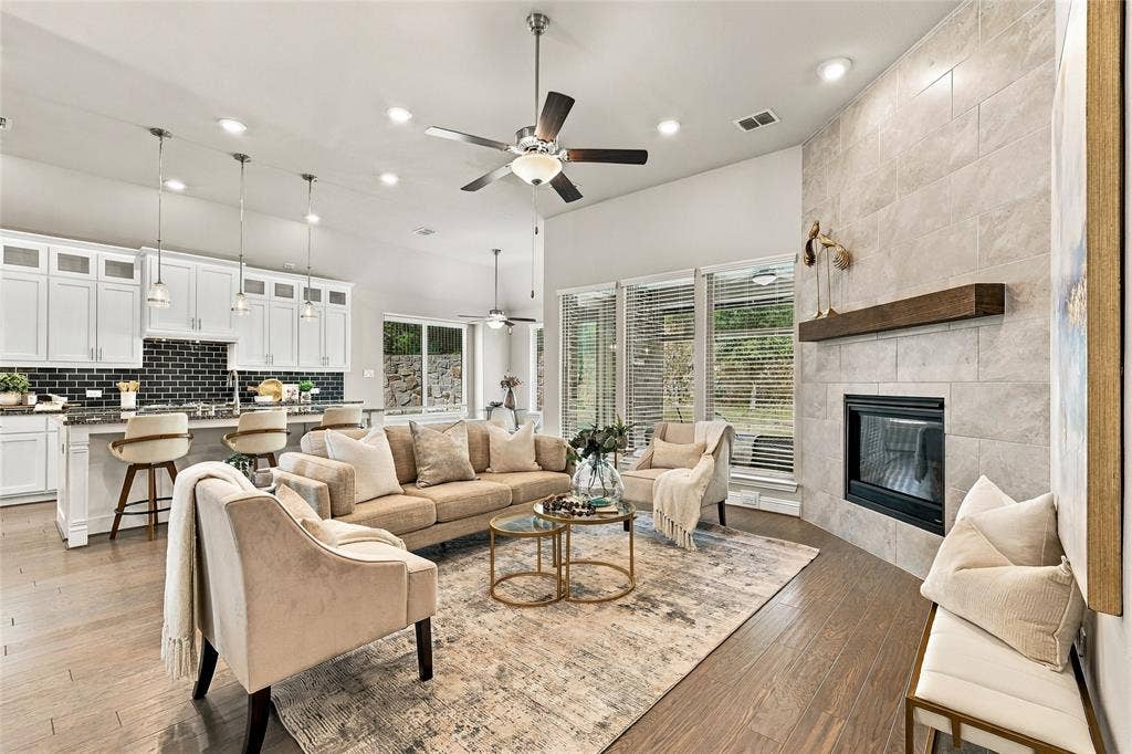 Open-concept interior featuring wood-finish flooring, a full-height stone-surround fireplace with a wooden mantel, white cabinetry, a kitchen island with pendant lighting, and recessed ceiling lights