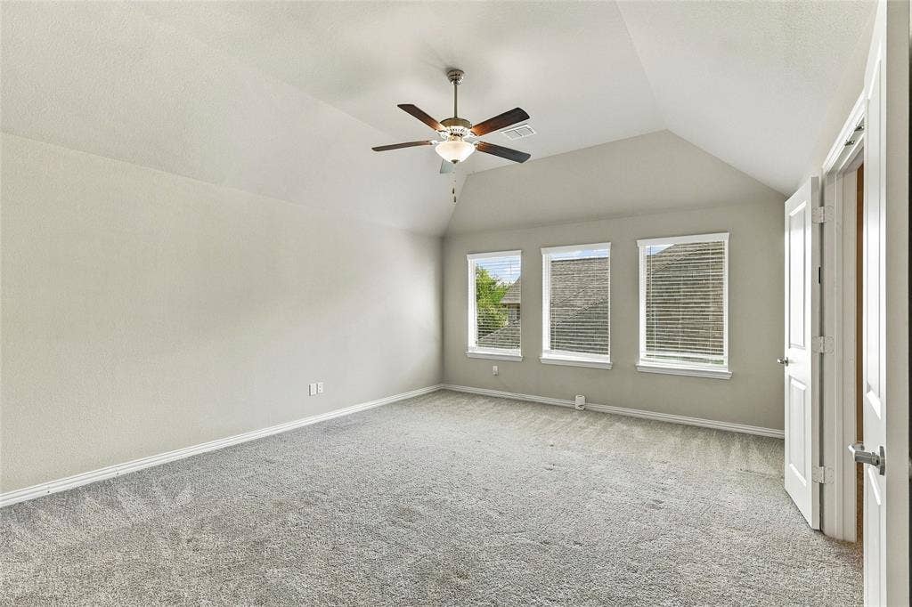 Vaulted ceiling featuring a central multi-blade ceiling fan with integrated light fixture, neutral wall palette, carpeting, and three windows