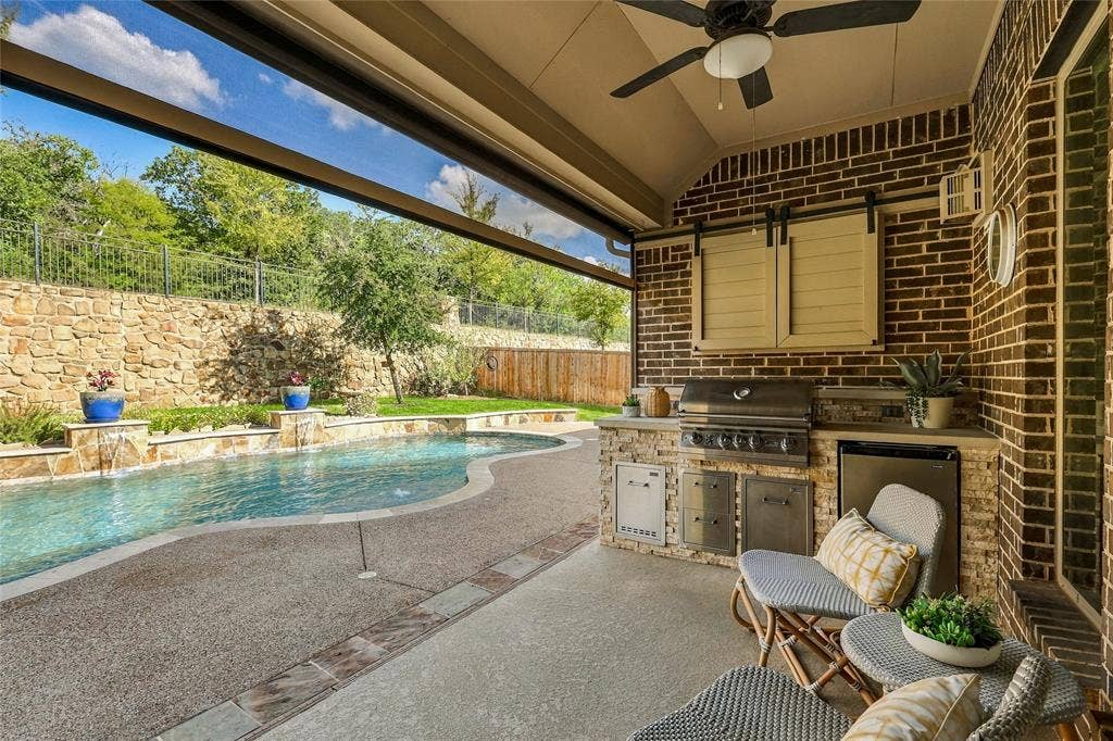 Covered patio featuring an outdoor kitchen with stainless steel appliances, stone masonry, a built-in ceiling fan, and a swimming pool with a stone retaining wall