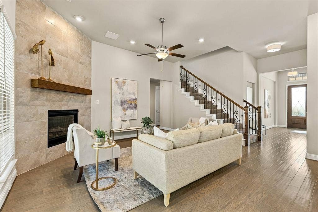 Floor-to-ceiling stone fireplace with a thick wood mantel, wood-finish flooring, staircase with ornate iron balusters, recessed lighting, and a ceiling fan with integrated light fixture