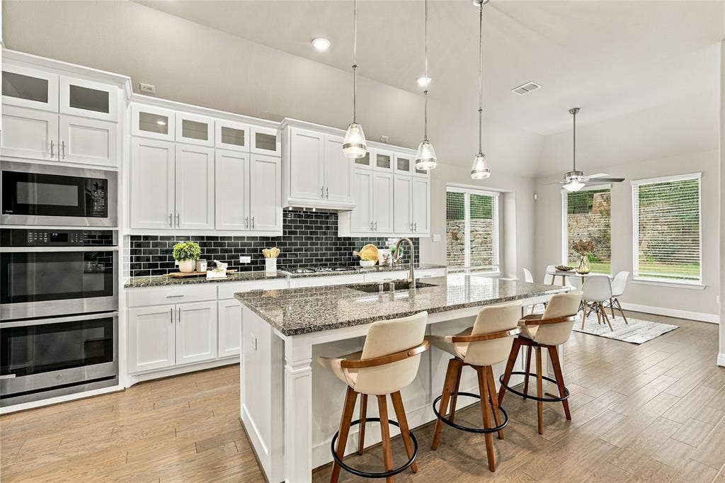 Kitchen featuring white cabinetry, stone countertops, a center island with a built-in sink, glass-tile backsplash, and wood-finish flooring