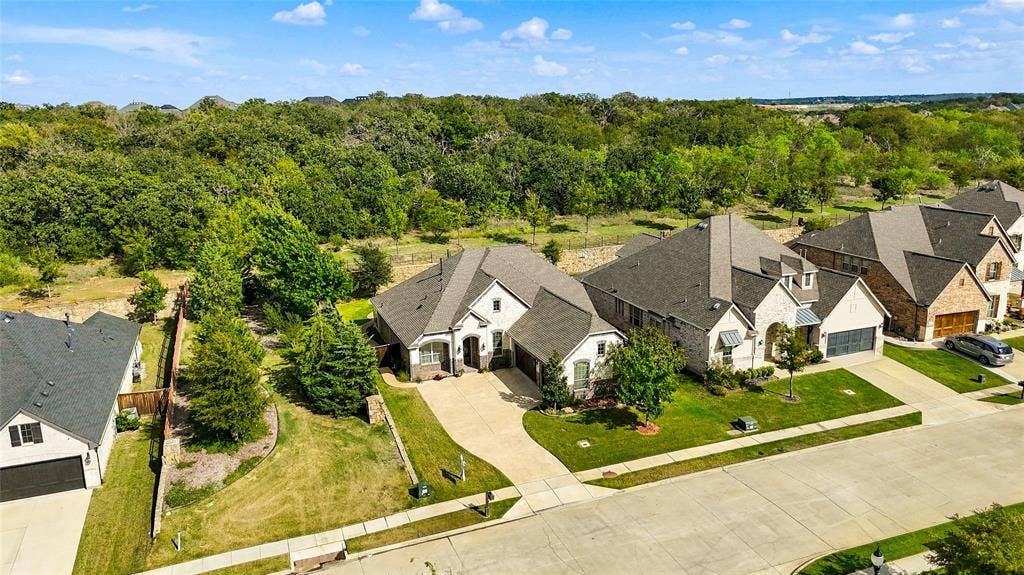 Aerial perspective featuring a multi-gabled brick and masonry exterior, asphalt shingle roofing, concrete driveway, and a perimeter fence line bordering an expanse of mature trees