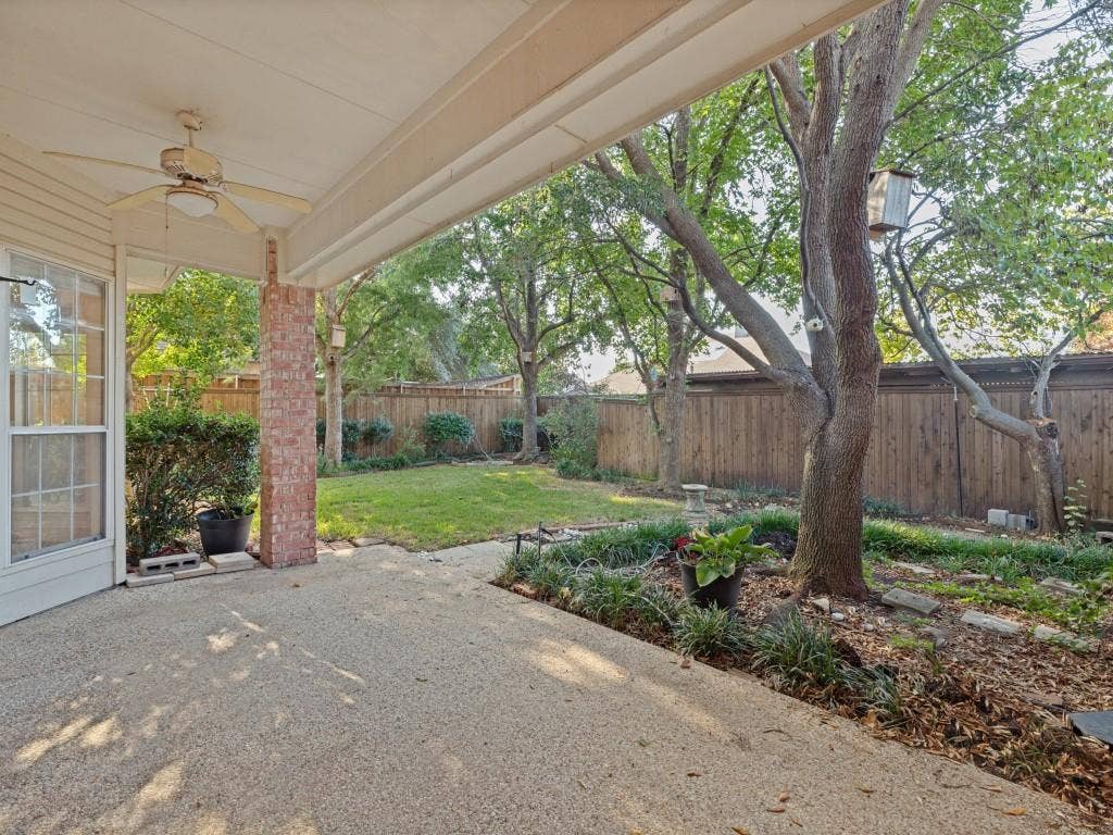 2817 Mayfair Lane, McKinney, TX 75071 Covered patio featuring an exposed aggregate concrete surface, brick support column, and overhead ceiling fan, opening to a fenced backyard with established trees