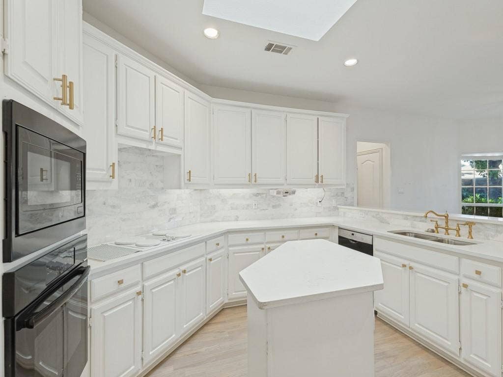 2817 Mayfair Lane, McKinney, TX 75071 Kitchen featuring white cabinetry with gold-tone hardware, stone-surface countertops, stone-tile backsplash, built-in dual wall ovens, and a center island under a skylight