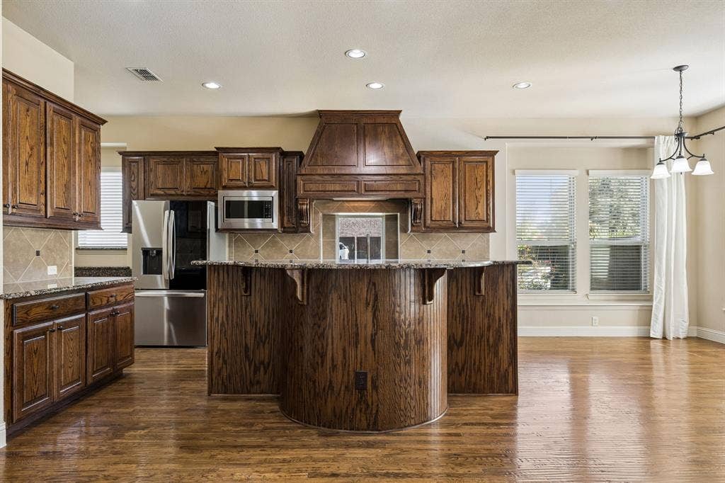 6448 Fire Creek Trail, Frisco, TX 75036 Kitchen featuring custom wood cabinetry, a curved island with stone countertops, stone tile backsplash, stainless steel appliances, and wood-finish flooring