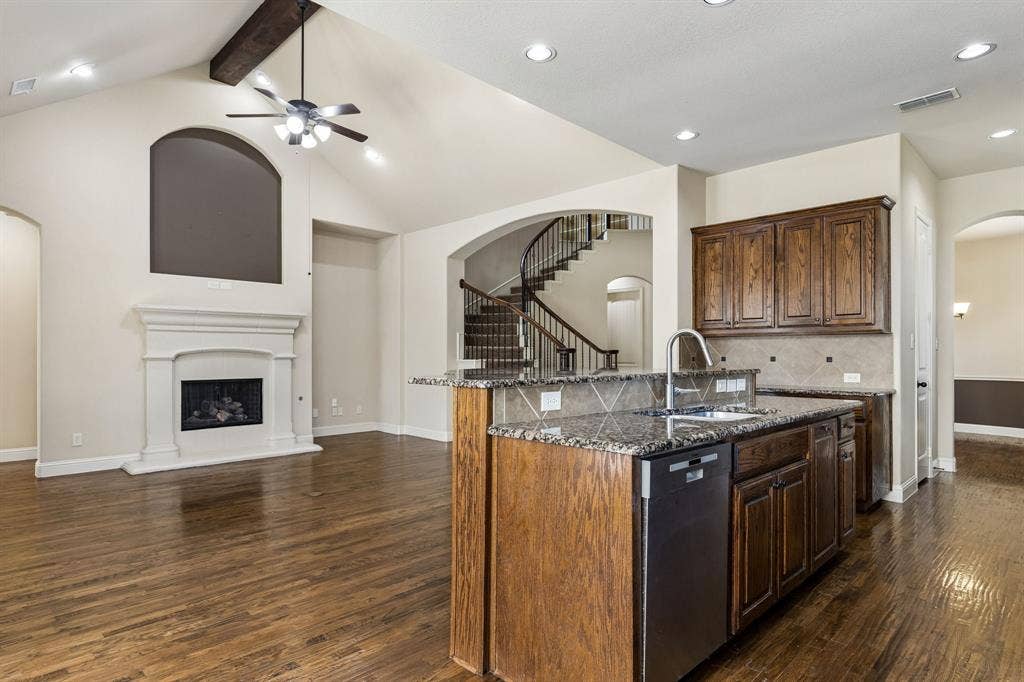 6448 Fire Creek Trail, Frisco, TX 75036 Vaulted ceilings with exposed timber beams, wood-finish flooring, masonry fireplace with decorative surround, kitchen island with stone-surface countertop, and curved staircase with wrought-iron railing