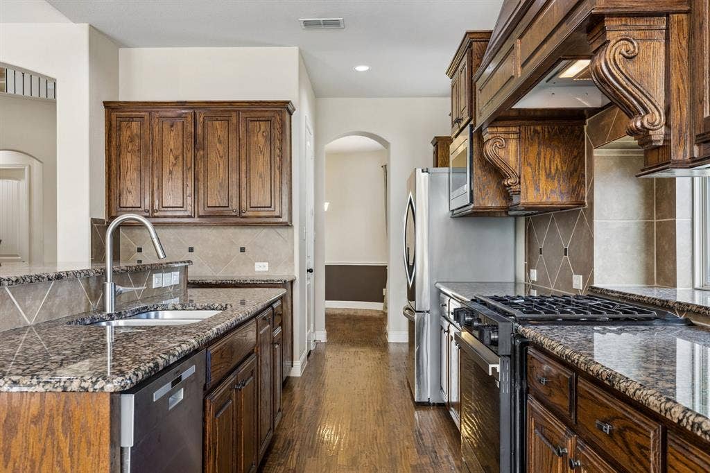 6448 Fire Creek Trail, Frisco, TX 75036 Kitchen featuring wood-finish cabinetry, stone countertops, a decorative range hood, stainless steel appliances, and diagonal tile backsplash