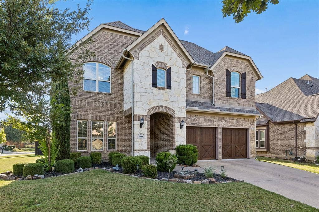 6448 Fire Creek Trail, Frisco, TX 75036 Two-story brick and stone veneer exterior featuring a covered arched entryway, decorative dark wood-finish carriage-style garage doors, dark shutters, and a multi-gabled roofline