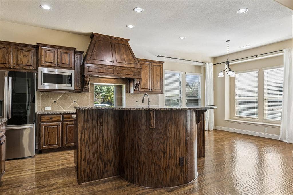 6448 Fire Creek Trail, Frisco, TX 75036 Kitchen featuring custom wood cabinetry, a curved island with stone countertops, a decorative wood range hood, tile backsplash, and wood-finish flooring