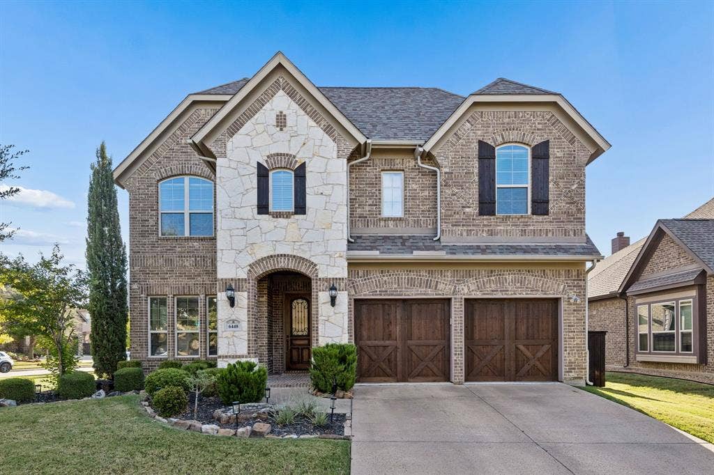 6448 Fire Creek Trail, Frisco, TX 75036 Two-story brick and stone facade with arched entryway, decorative wood-finish shutters, double carriage-style garage doors, and arched window detailing