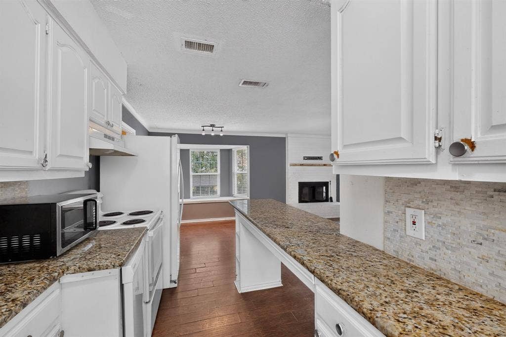 9651 Cherry Street, Frisco, TX 75033 Kitchen featuring white cabinetry, stone-finish countertops, tile backsplash, and wood-finish flooring, extending toward a fireplace with a mantel