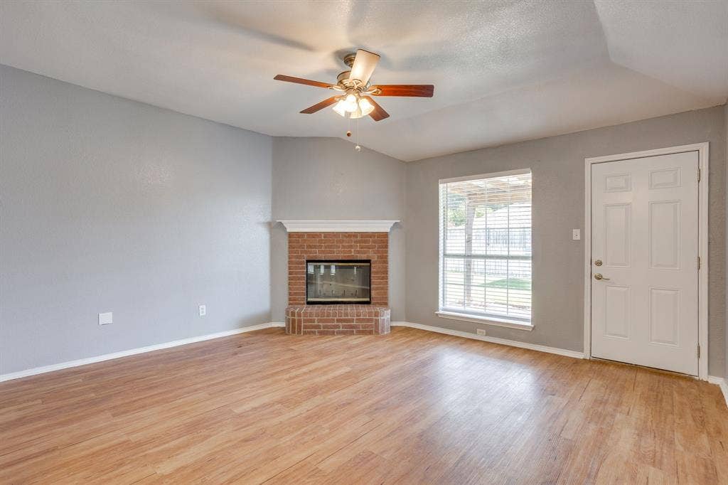 6221 St Leonard Drive, Arlington, TX 76001 Vaulted ceiling featuring a ceiling fan with integrated lighting, wood-finish flooring, corner brick fireplace with a mantel, and a single-light exterior door