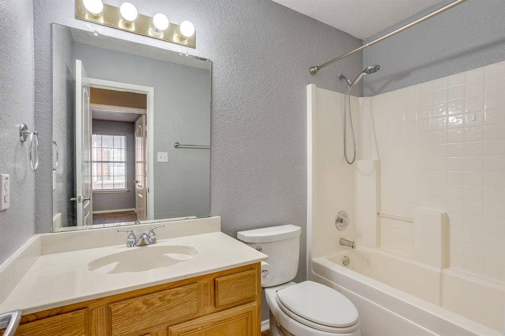 6221 St Leonard Drive, Arlington, TX 76001 Bathroom featuring a wood-finish vanity with a seamless integrated countertop and sink, wall-mounted four-light fixture, large rectangular vanity mirror, and a tub-shower combination with a showerhead attachment