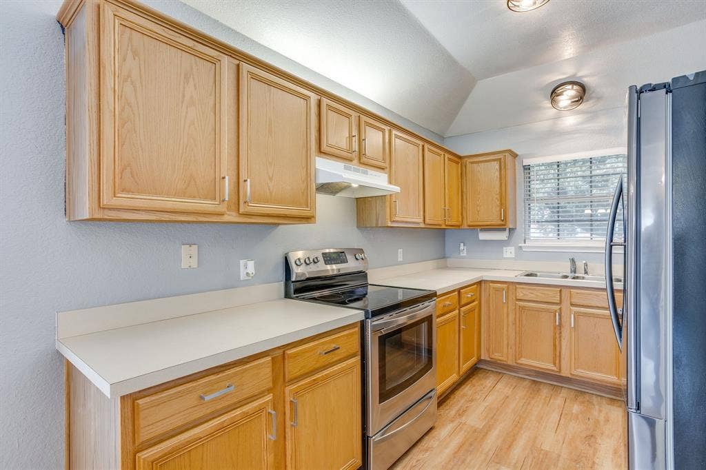 6221 St Leonard Drive, Arlington, TX 76001 Kitchen featuring wood cabinetry, stainless steel appliances, solid-surface countertops, wood-finish flooring, and a vaulted ceiling