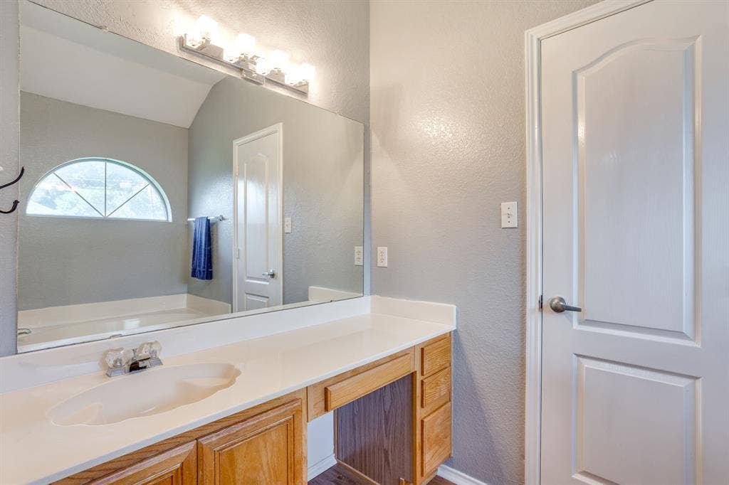 6221 St Leonard Drive, Arlington, TX 76001 Bathroom vanity with a solid-surface countertop, integrated sink, wood-finish cabinetry, a large wall-mounted mirror, and an arched window