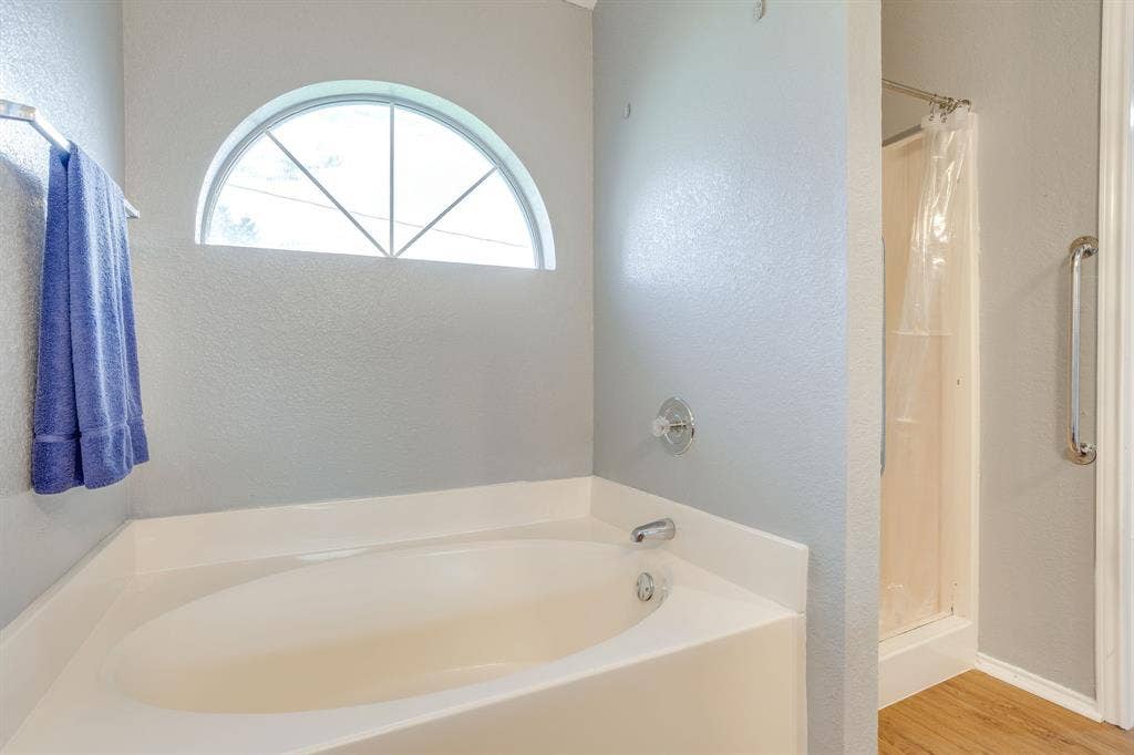 6221 St Leonard Drive, Arlington, TX 76001 Oval soaking tub with integrated surround, arched window, wood-finish flooring, and shower enclosure with a wall-mounted grab bar