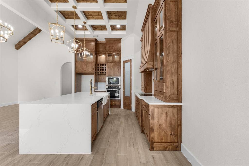 Kitchen featuring custom stained-wood cabinetry, a large waterfall island with quartz countertops, a coffered ceiling with wood-finish inlays, integrated stainless steel double ovens, and wood-finish flooring