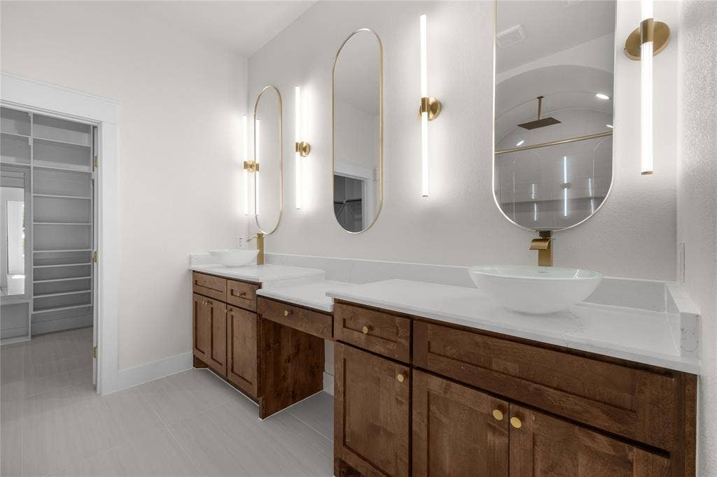 Dual wood cabinetry vanity with white countertops, vessel sinks, gold-tone hardware, vertical wall-mounted lighting, and arched mirrors