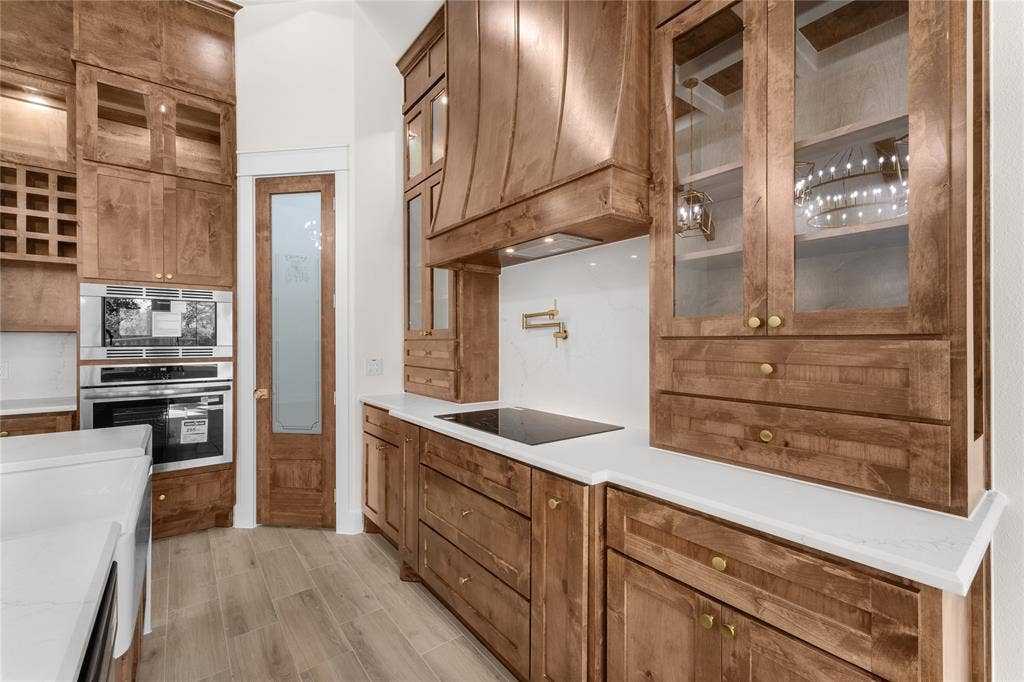Custom wood cabinetry featuring glass-front display uppers and an integrated wine rack, complemented by stone-finish countertops, a matching wood-paneled range hood, and a gold-finish pot filler faucet