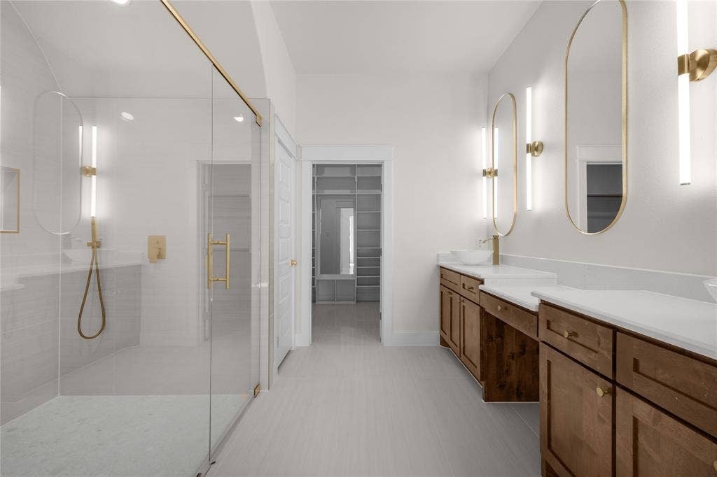 Glass-enclosed walk-in shower with gold-finish hardware, dual vanity with solid-surface countertops, vessel sinks, oval-framed wall mirrors, and integrated shelving in the dressing area