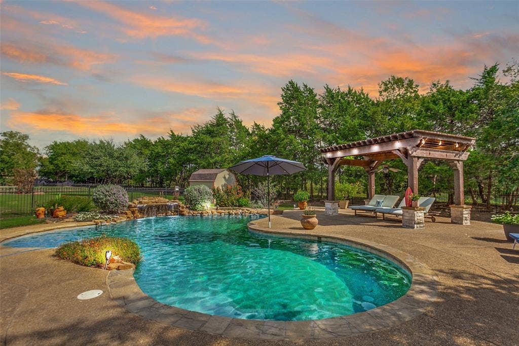 214 Stanford Ct. - Resort-style pool with waterfall feature and diving rock, surrounded by beautiful landscaping, pergola, and ample space for outdoor entertaining and sunbathing. AI generated sunset & ligting
