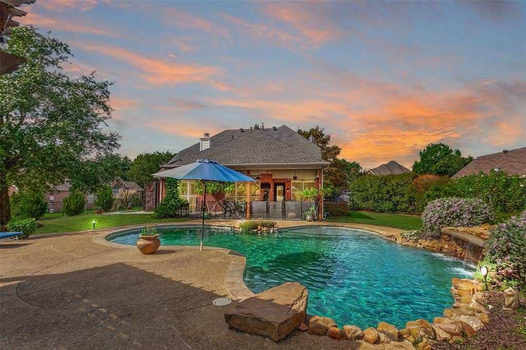 214 Stanford Ct. - Resort-style pool with waterfall feature and diving rock, surrounded by beautiful landscaping, pergola, and ample space for outdoor entertaining and sunbathing. AI generated sunset.