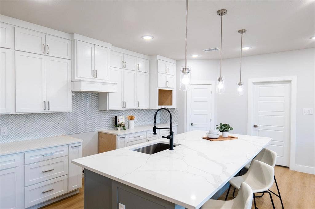 Kitchen featuring white shaker-style cabinetry, quartz countertops with a veined pattern, herringbone-patterned backsplash tile, center island with an undermount sink and matte black faucet, and three pendant lights