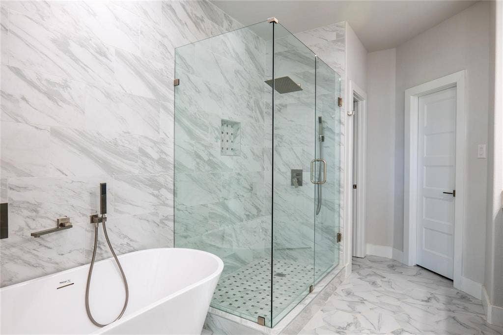 Freestanding soaking tub with wall-mounted hand shower, glass-enclosed shower with tiled walls, rainfall showerhead, and patterned tile flooring