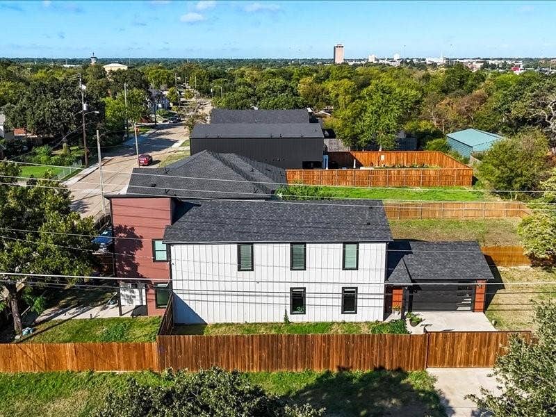 Multi-story modern residence featuring vertical panel siding, dark-toned shingle roofing, integrated garage with horizontal panel door, and a perimeter privacy fence enclosing the lot