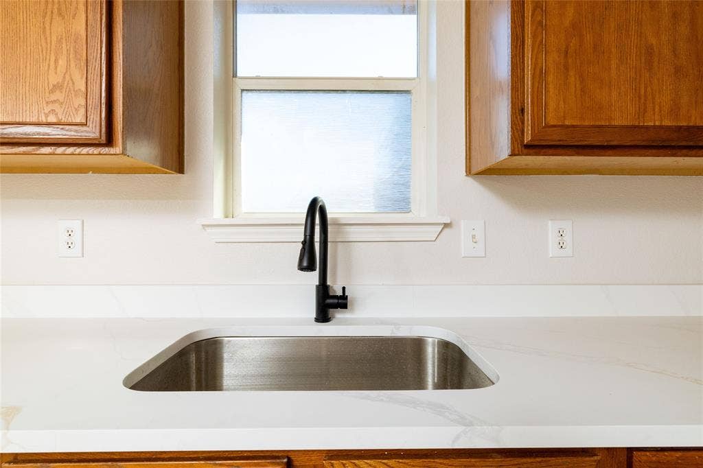 Undermount stainless steel kitchen sink featuring a matte black gooseneck faucet, white composite countertop with subtle veining, and natural-finish wood cabinetry