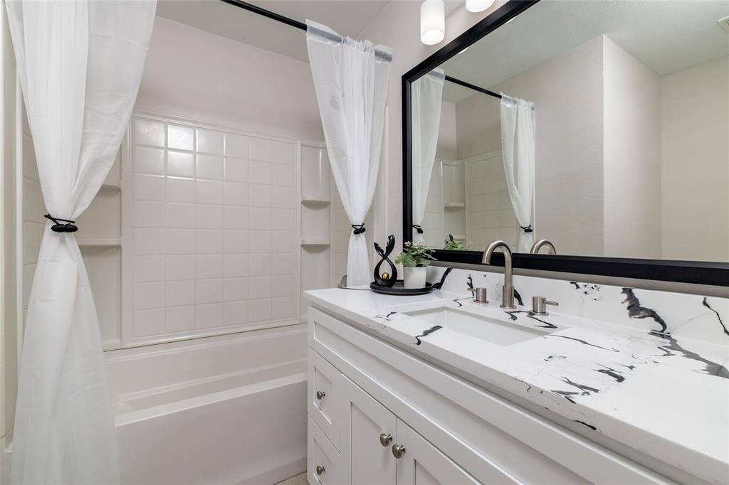 Bathroom featuring a white vanity with a patterned stone countertop, integrated sink, matte black-framed mirror, and a tub-shower combination with a tiled surround